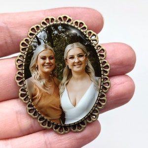 Pu&ograve; includere: Una cornice per foto ovale in bronzo con un bordo a festoni. La cornice contiene una foto di due donne che sorridono alla telecamera. Le donne indossano abiti casual e sono in piedi in una zona boscosa.