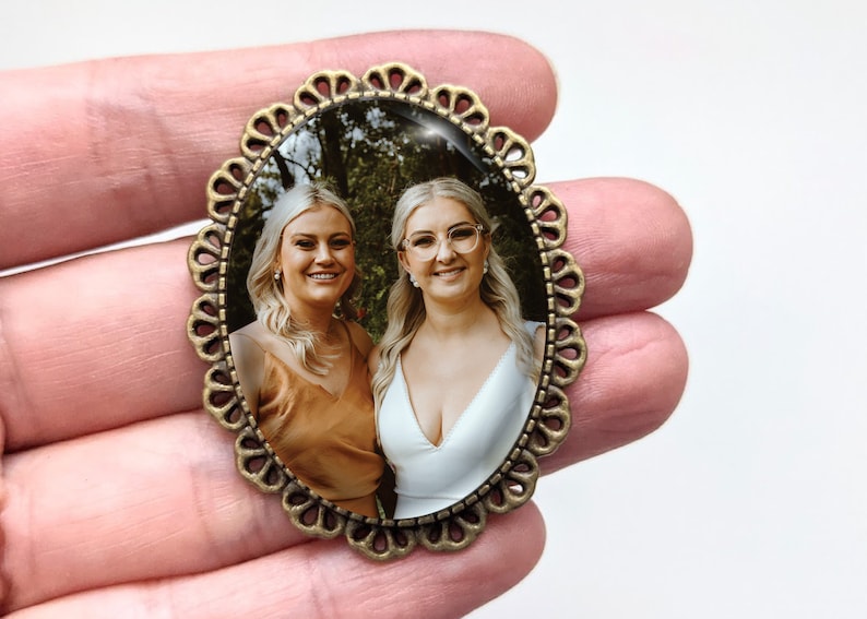 Pu&ograve; includere: Una cornice per foto ovale in bronzo con un bordo a festoni. La cornice contiene una foto di due donne che sorridono alla telecamera. Le donne indossano abiti casual e sono in piedi in una zona boscosa.
