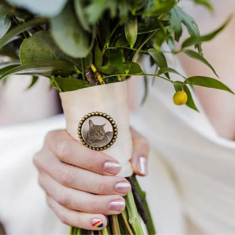 May include: A white bridal bouquet with a round, gold-toned charm featuring a photo of a cat. The charm is attached to the bouquet with a white ribbon.