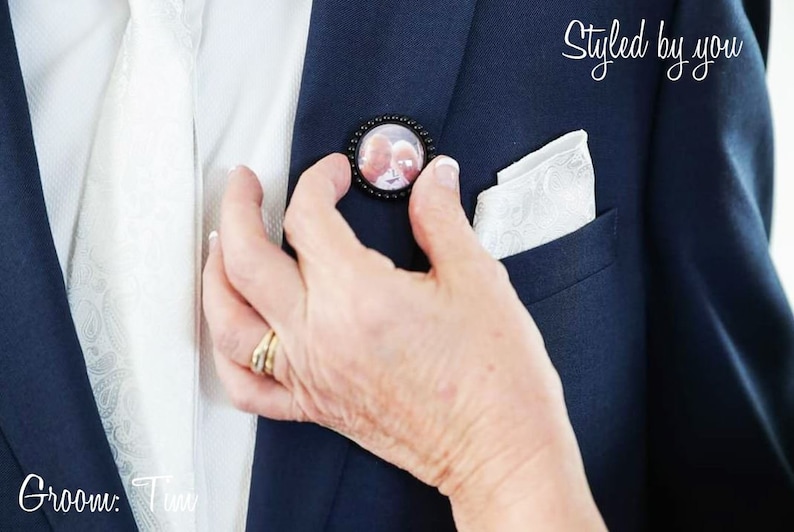 May include: A person adjusting a black button with a photo inside on a navy blue suit jacket. The jacket has a white pocket square. The text "Styled by you" is visible in the upper right corner of the image.