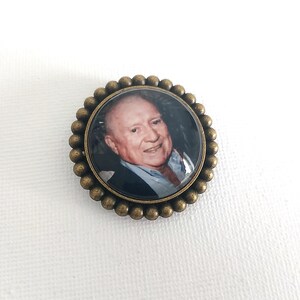 May include: A bronze-colored round pin with a photo of a man in a dark suit and a light blue shirt. The man is smiling and looking at the camera.