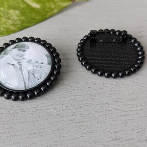 May include: Black and white photo of a couple in a round glass photo charm with a black beaded border. The back of the charm is black with a pin.