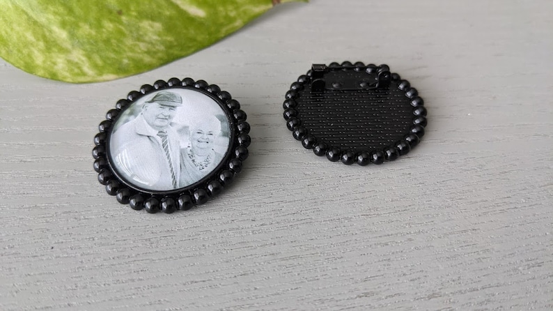 May include: Black and white photo of a couple in a round glass photo charm with a black beaded border. The back of the charm is black with a pin.