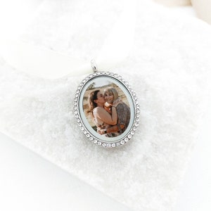 May include: Silver oval locket with a clear crystal border. The locket contains a photo of two people hugging.