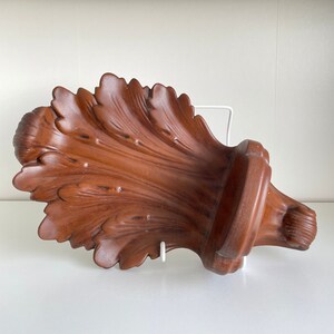May include: A brown, leaf-shaped wall bracket with a carved, textured surface. The object features a wide, fan-like arrangement of foliage patterns that taper towards a rounded, solid base, creating a decorative shelf-like structure for mounting.