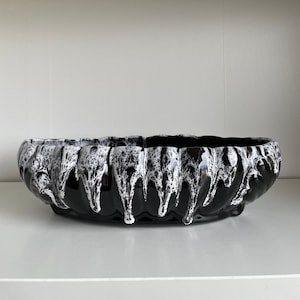May include: A black ceramic bowl with a wavy rim and a glossy finish. The bowl features a white, dripped glaze design around the rim, creating a contrasting texture. The bowl is a decorative piece.