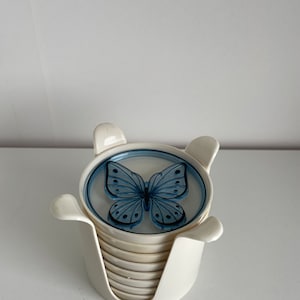 May include: A set of white ceramic coasters with a blue butterfly design. The coasters are stacked in a holder.