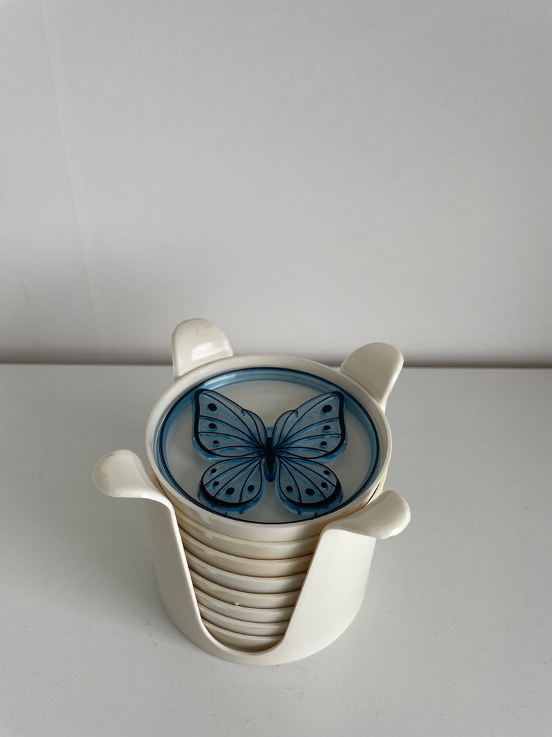 May include: A set of white ceramic coasters with a blue butterfly design. The coasters are stacked in a holder.