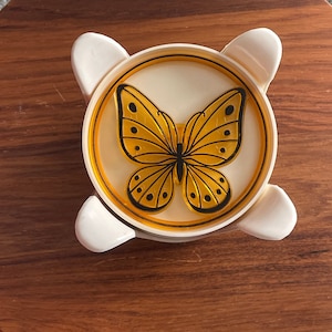May include: A white plastic butterfly-shaped dish with a yellow butterfly design. The dish has a raised rim and a smooth surface.