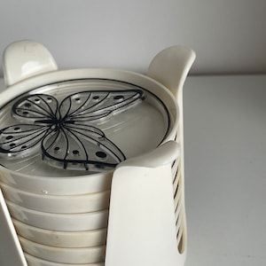 May include: Set of six white coasters with a black butterfly design. The coasters are stacked in a white holder.