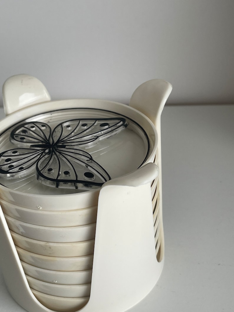 May include: Set of six white coasters with a black butterfly design. The coasters are stacked in a white holder.