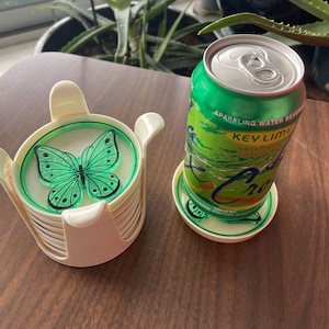 May include: A set of six white coasters with a green butterfly design and a green can of sparkling water beverage with the text "Key Lime Natural Essence" and "Cran" on the label.