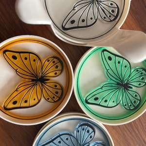 May include: Set of four white ceramic coasters with a butterfly design. Each coaster features a different colored butterfly: orange, green, blue, and white. The butterflies have black outlines and black dots on their wings.