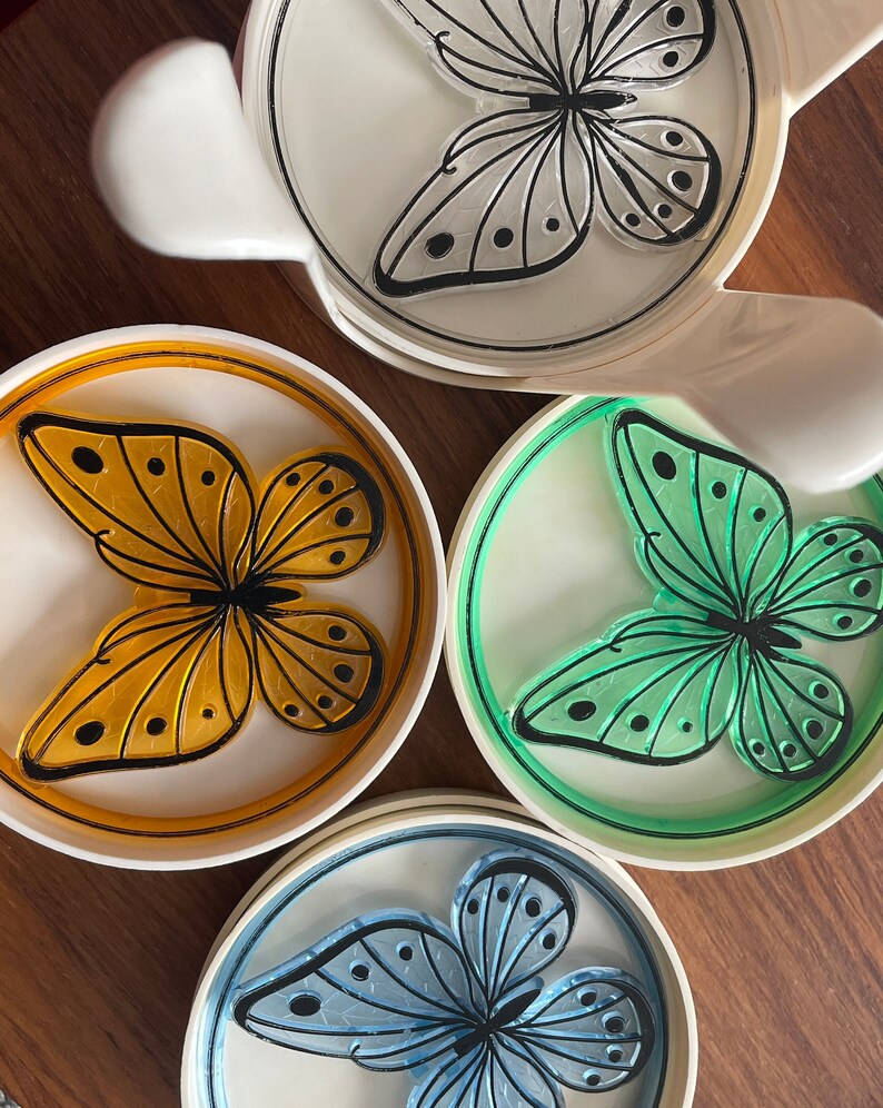 May include: Set of four white ceramic coasters with a butterfly design. Each coaster features a different colored butterfly: orange, green, blue, and white. The butterflies have black outlines and black dots on their wings.