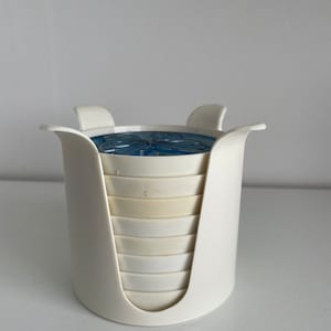 May include: White plastic coaster holder with a stack of white coasters with a blue and white design on the top coaster.