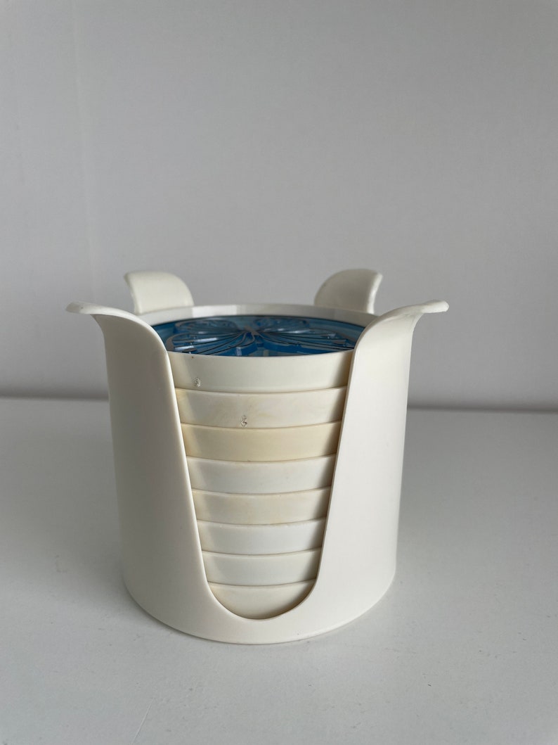 May include: White plastic coaster holder with a stack of white coasters with a blue and white design on the top coaster.