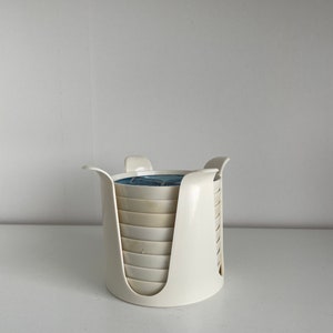 May include: A set of white ceramic coasters with a blue and white design, stacked in a white plastic holder.