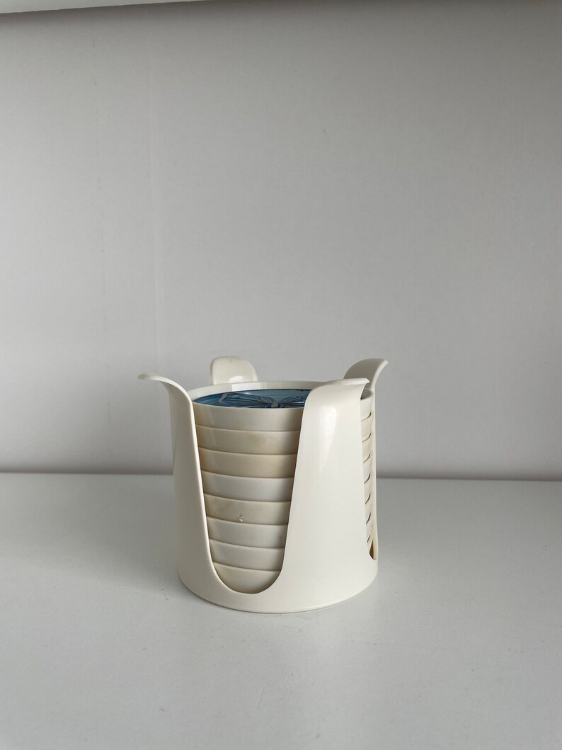 May include: A set of white ceramic coasters with a blue and white design, stacked in a white plastic holder.