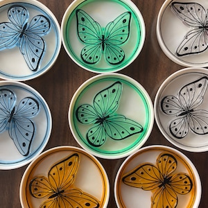 May include: Set of six round coasters featuring a butterfly design. Each coaster has a white background with a different colored butterfly in the center. The butterflies are green, blue, orange, and gray.