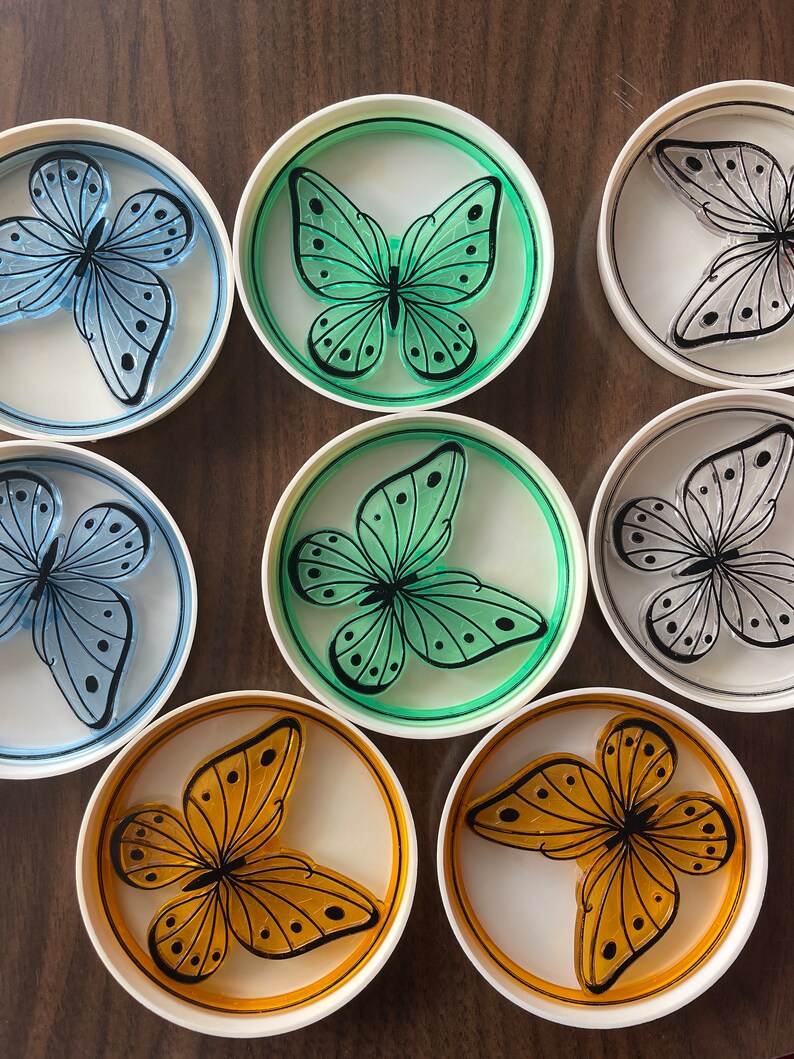 May include: Set of six round coasters featuring a butterfly design. Each coaster has a white background with a different colored butterfly in the center. The butterflies are green, blue, orange, and gray.