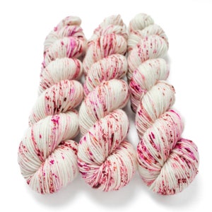 May include: Three skeins of white yarn with pink and red speckles. The yarn is arranged in a loose, twisted formation.