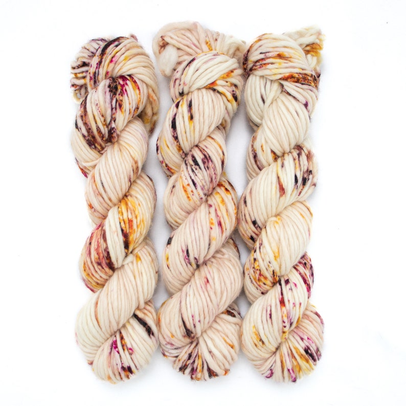 May include: Three skeins of yarn in a cream colour with flecks of burgundy, orange, and yellow.