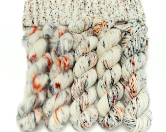 May include: A collection of five skeins of yarn in various textures and weights, with a speckled headband. The yarn colors are primarily cream with flecks of orange, black, and gray. The labels read: Dandelion Mohair, Alpaca Floof, Staple Sock, Super Squishy Worsted, and Maizy Super Bulky.