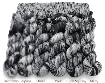 May include: Six skeins of black and white variegated yarn arranged in a row, labelled Dandelion Mohair, Alpaca Floof, Staple Sock, Pixie DK, Super Squishy Worsted, and Maizy Super Bulky, with a textured black and white knit fabric swatch above.