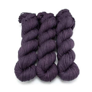 May include: Three skeins of dark purple yarn are arranged side by side against a white background. The yarn is tightly wound into a compact, cylindrical shape, with a slightly textured surface. The color is a deep, saturated shade of purple.