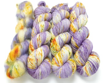 May include: Four skeins of yarn in shades of purple, white, and yellow. The yarn is arranged in a row, with the skeins on the left and right being slightly smaller than the two in the middle.
