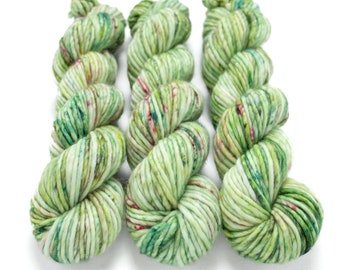 May include: Three skeins of yarn in a green, white, and pink colorway. The yarn is a chunky weight and has a speckled appearance.