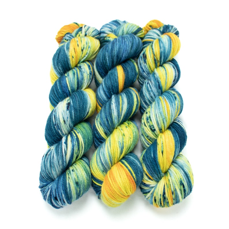 Op de afbeelding: Drie bollen garen in een blauwe, gele en groene kleurstelling. Het garen is een gem&ecirc;leerde kleurstelling met een gespikkeld effect.
