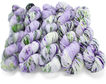 May include: A set of six skeins of yarn in shades of purple, green, and white. The yarn is hand-dyed and has a speckled appearance.
