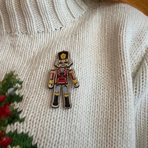 Peut inclure: Une décoration de casse-noisette en bois avec un motif peint rouge et jaune. Le casse-noisette porte un chapeau bleu et or et a une moustache. La décoration est épinglée sur un pull blanc.