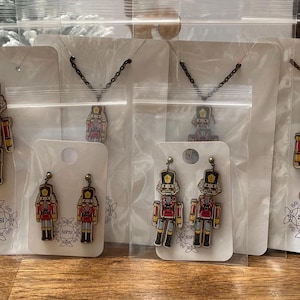 Puede incluir: Cuatro juegos de joyas con temática de cascanueces, que incluyen collares y pendientes. Cada juego presenta una figurita de cascanueces roja y dorada con un sombrero negro y detalles blancos. Las joyas se muestran en tarjetas blancas con la palabra "SIPSI" impresa en ellas.