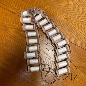 May include: A collection of vintage wooden spools, each wound with white thread, arranged in a chain-like pattern. The spools are connected by black thread, creating a unique decorative element. The wooden spools have a natural wood grain finish.