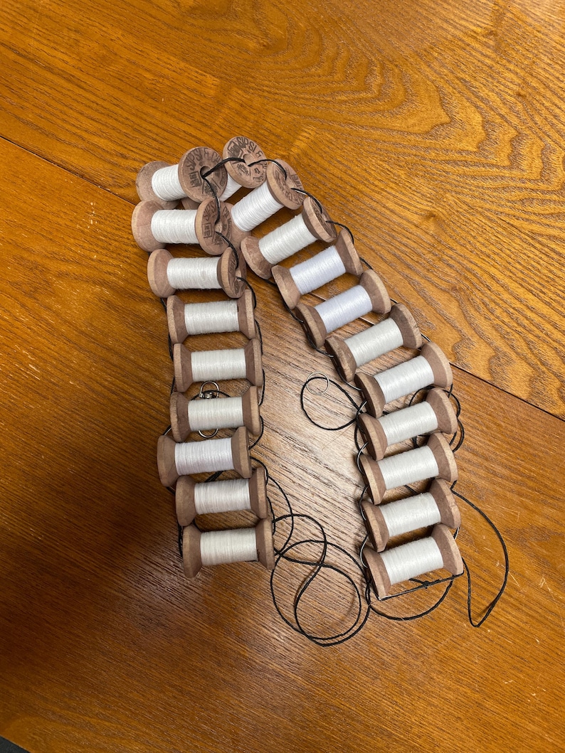 May include: A collection of vintage wooden spools, each wound with white thread, arranged in a chain-like pattern. The spools are connected by black thread, creating a unique decorative element. The wooden spools have a natural wood grain finish.