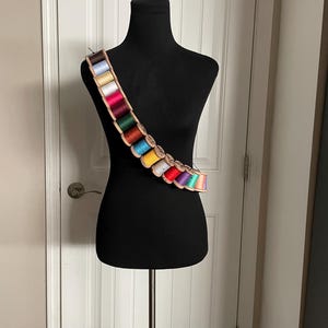 May include: A black dress form displays a bandolier-style thread holder. The holder is made of light brown wood and holds spools of colorful thread in various shades of blue, green, red, yellow, and purple. The dress form is on a metal stand.