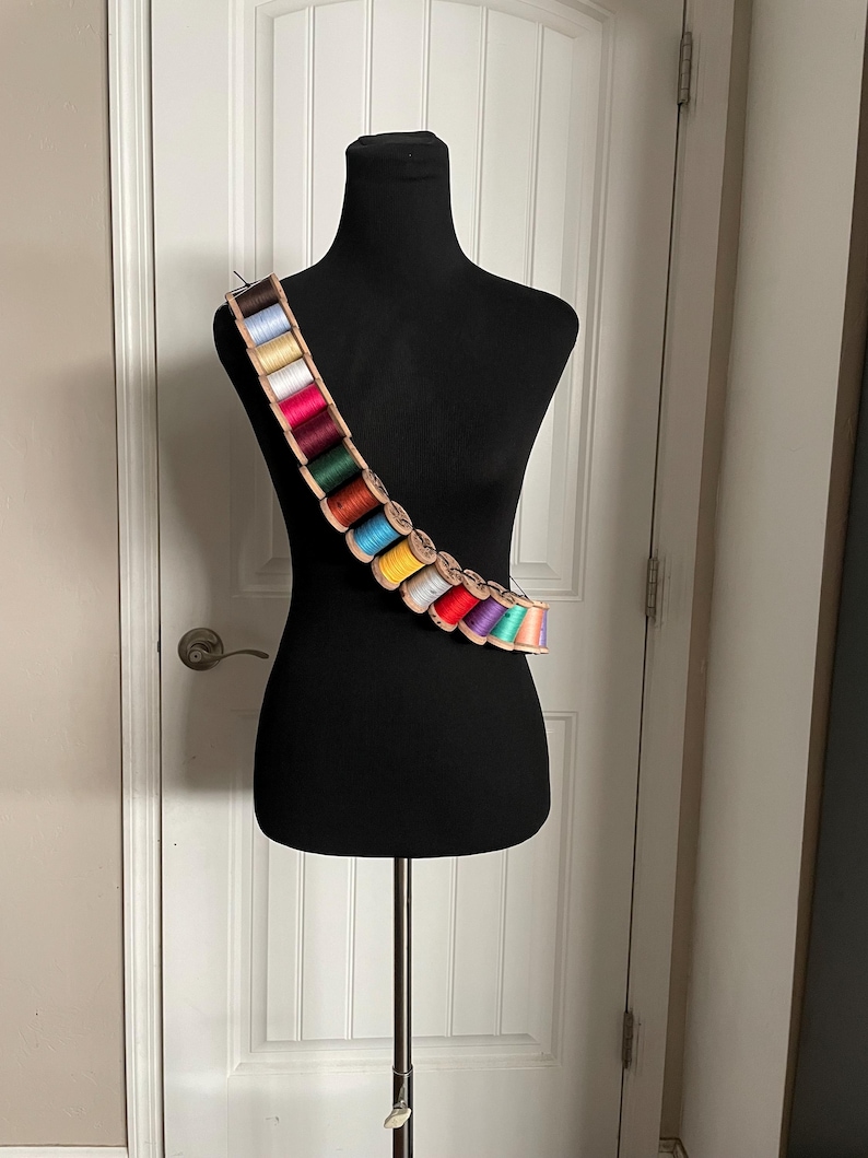 May include: A black dress form displays a bandolier-style thread holder. The holder is made of light brown wood and holds spools of colorful thread in various shades of blue, green, red, yellow, and purple. The dress form is on a metal stand.