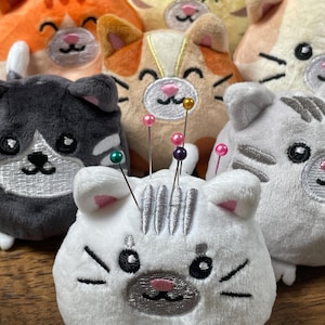 May include: A collection of plush cat-shaped pin cushions in various colors, including orange, gray, and white. Each cushion has embroidered facial features and is adorned with colorful pins, perfect for sewing enthusiasts.