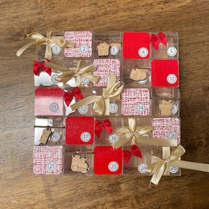 Könnte beinhalten: Ein roter und weißer Weihnachts-Adventskalender mit 25 durchsichtigen Plastikboxen, die jeweils einen kleinen Holzschmuck enthalten. Die Boxen sind in einem 5x5-Raster angeordnet und mit rotem und goldenem Band gebunden.