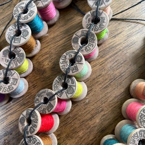May include: A close-up of a handmade garland composed of vintage wooden spools. Each spool is filled with colorful thread in shades of pink, red, blue, yellow, and brown. The spools are strung together with black thread, creating a decorative craft item.
