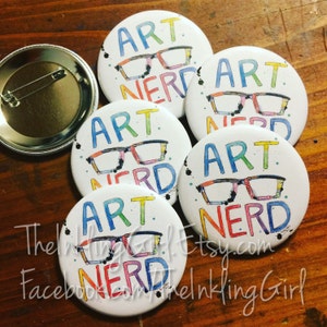 May include: Five white round buttons with the text "ART NERD" in rainbow colors and a pair of glasses made of colorful pencils. The text is in a handwritten style.