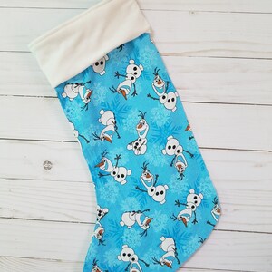 May include: A blue Christmas stocking with a white cuff. The stocking features a repeating pattern of Olaf from the movie Frozen. The pattern includes Olaf's face, body, and arms. The background is a light blue with white snowflakes.