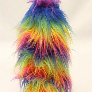 May include: A tall, shaggy, multi-colored faux fur item. The colors include purple, blue, green, yellow, orange, and pink. The item has a layered appearance with long, wispy strands of fur extending outwards.