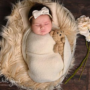 May include: A newborn infant wrapped in a cream-colored swaddle blanket, wearing a cream bow headband, resting in a round basket lined with cream faux fur. A small, light brown crocheted teddy bear is tucked into the swaddle next to the infant.