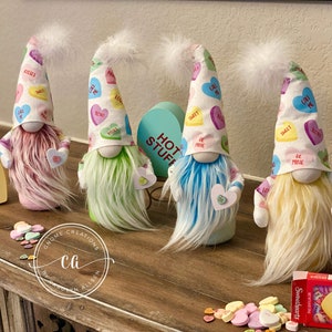 May include: Four Valentine's Day gnomes with candy heart patterned hats and fuzzy white beards. The gnomes are standing on a wooden table with a box that says "Hot Stuff" and another box that says "OYOX". There are also a few scattered candy hearts on the table.