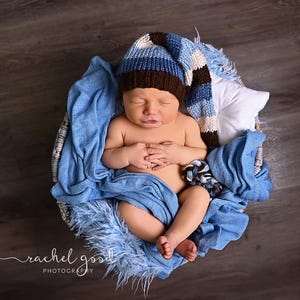 May include: A newborn baby sleeping in a round basket lined with a blue textured fabric. The baby wears a long, striped knit cap featuring bands of brown, light blue, and white. A small white pillow is tucked behind the baby's head in the basket.