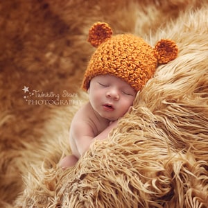Könnte beinhalten: Ein Baby trägt eine braune Häkelmütze mit Bärenohren und liegt auf einer braunen, flauschigen Decke.