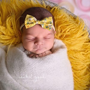 May include: A newborn infant wrapped in a soft white blanket, resting in a basket lined with bright yellow faux fur. The infant wears a yellow headband featuring a small bow with a floral and animal print pattern, eyes closed in sleep.